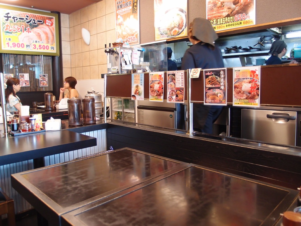 View of a bustling ramen shop interior with customers and a chef preparing a bowl
